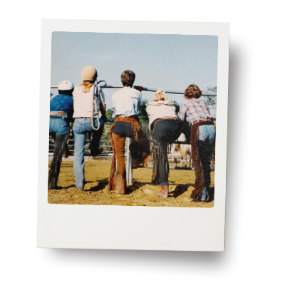 Five cowgirls facing away from us, leaning on the fence watching horses.