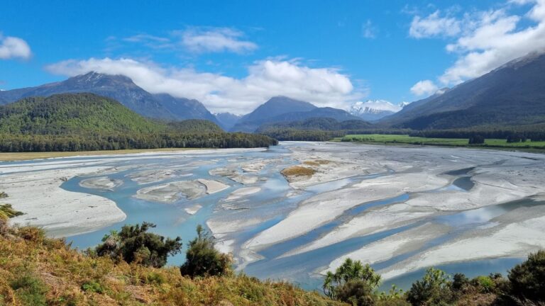 A wide, peaceful view of the Dart River Valley in South Island, New Zealand, with braided river channels, forest, and distant mountains under a bright blue sky.