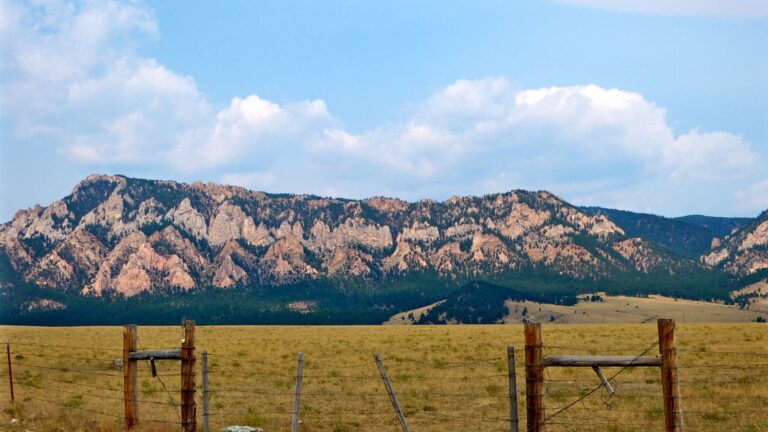 Wide open California backcountry with a wooden fence in the foreground, dry grassland, and rugged mountain ridges under a soft blue sky