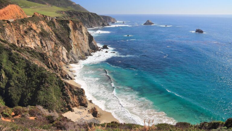 Rocky California coastline with steep cliffs, turquoise ocean water, and waves breaking along a quiet, open shoreline under a clear sky
