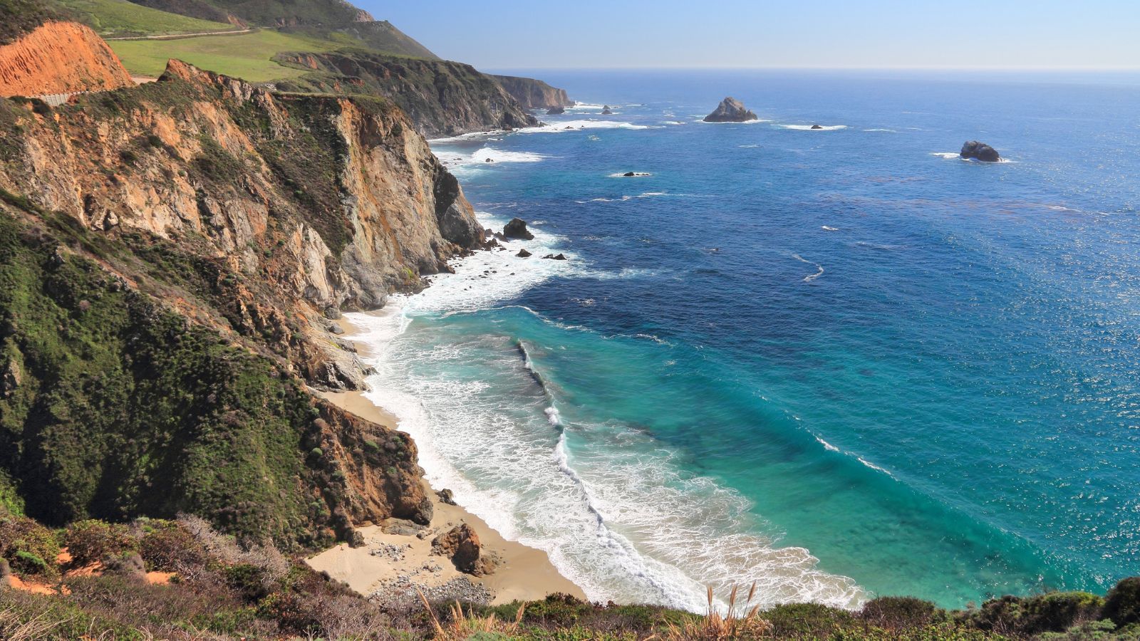 Rocky California coastline with steep cliffs, turquoise ocean water, and waves breaking along a quiet, open shoreline under a clear sky
