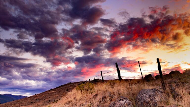 Dramatic sunset clouds over dry hills and fence line near Queenstown, Aotearoa New Zealand, with warm light breaking through the sky